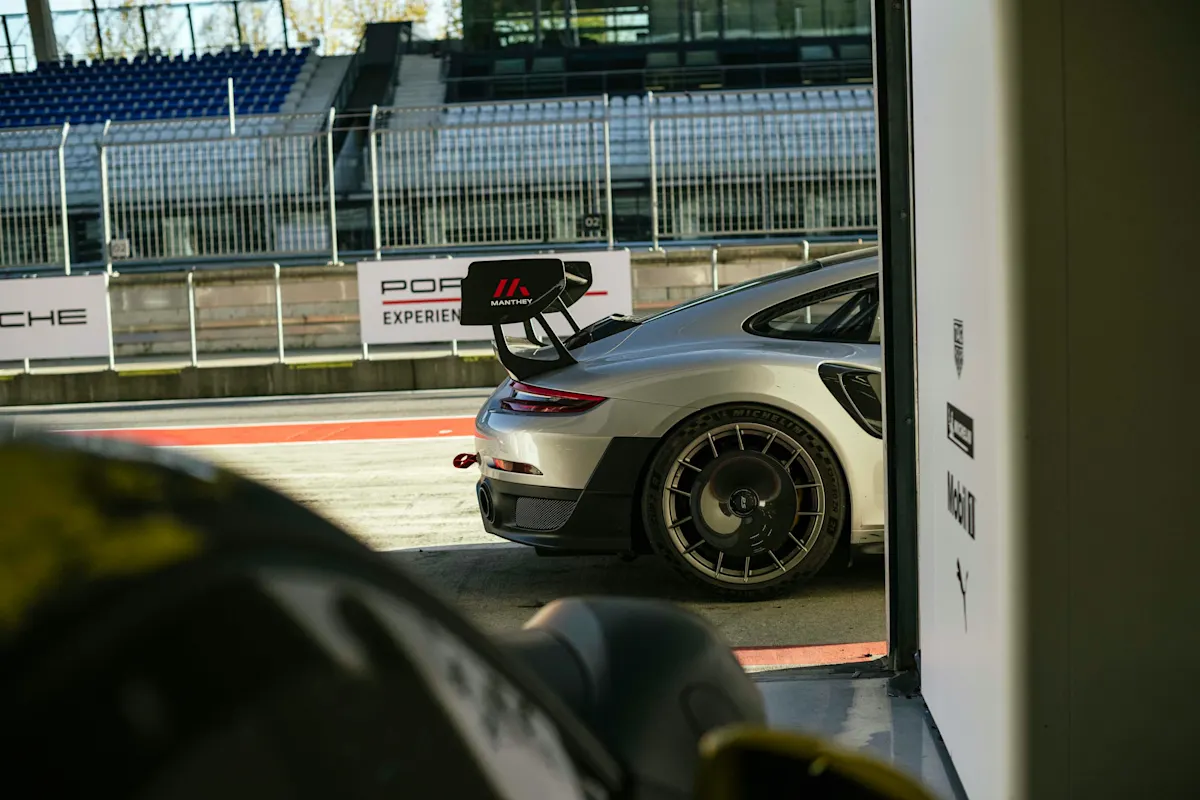 Porsche Rennwagen mit Heckspoiler am Eingang der Boxengasse, im Hintergrund Tribünen und Schilder.