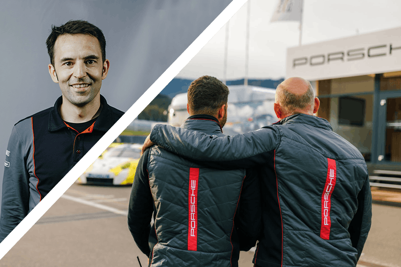 Man in Porsche jacket, smiling with friend, watching racetrack.