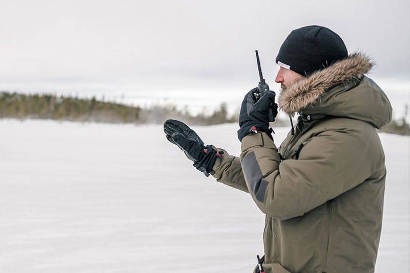 Person in winter gear using a walkie-talkie in snowy landscape with trees in distance.