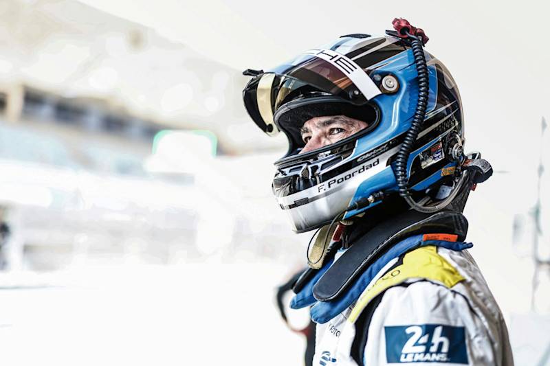 Porsche race car driver in full gear and helmet with attached headset, standing in profile view.
