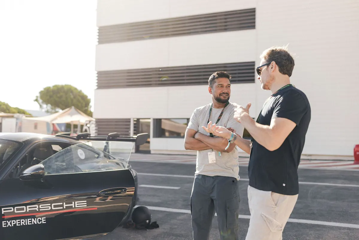 Two people chatting by a black Porsche car outside a building on a sunny day.