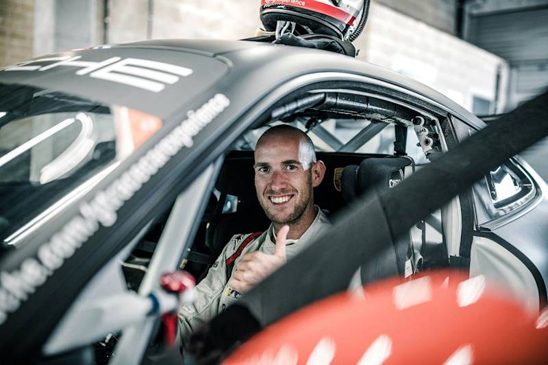 Person in racing suit inside Porsche race car, giving thumbs up with helmet on roof.