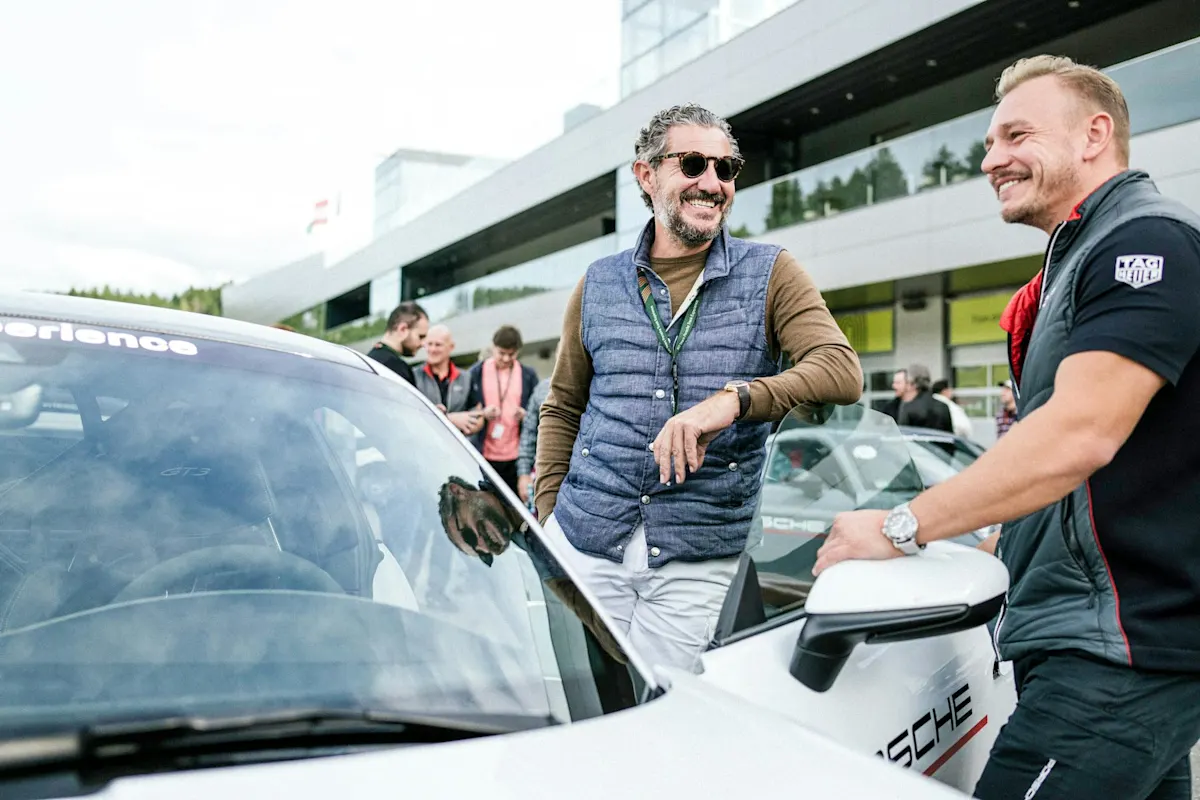 Two men smiling next to a white Porsche, with people and a building in the background.