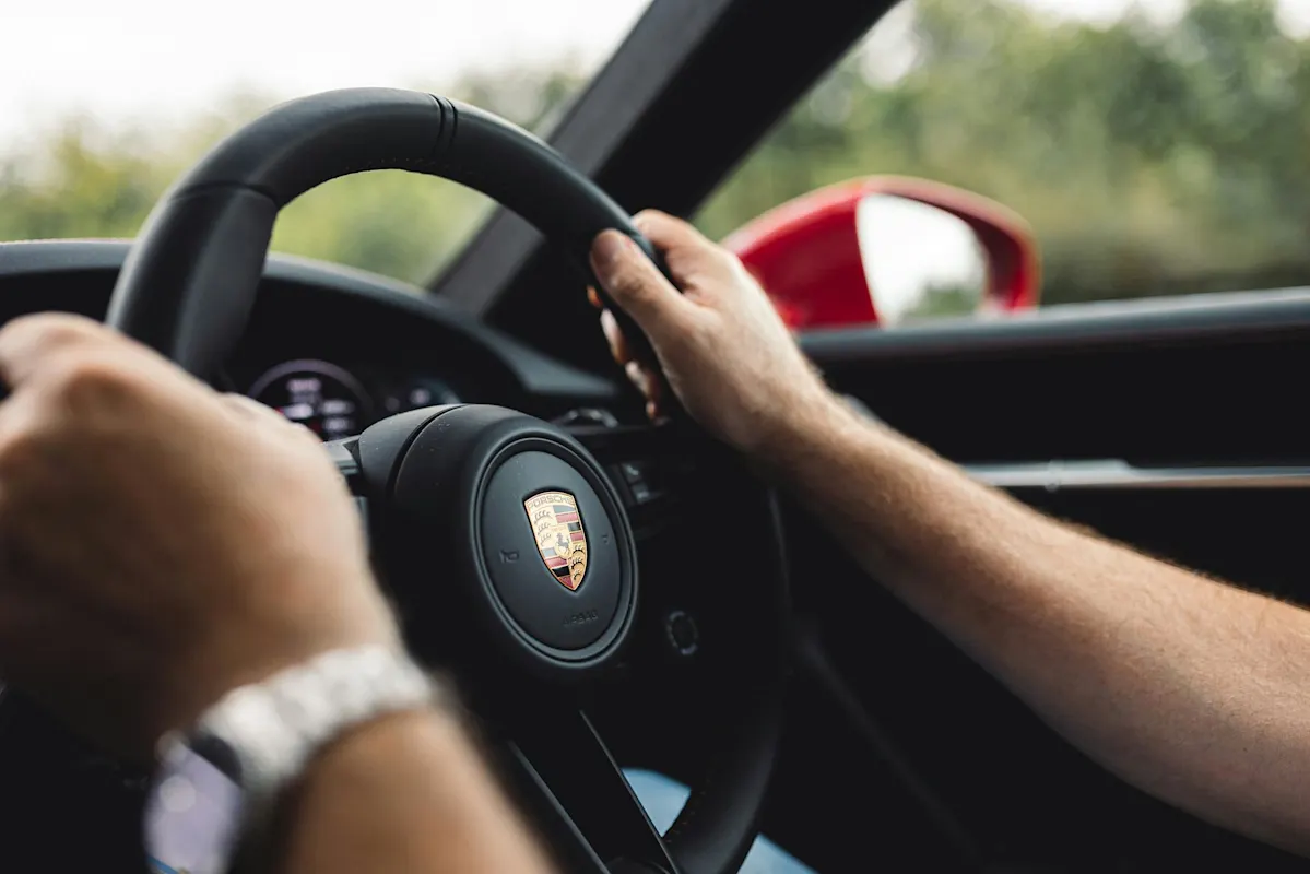 Person driving a Porsche car with logo visible on the steering wheel. Blurred red side mirror in view.