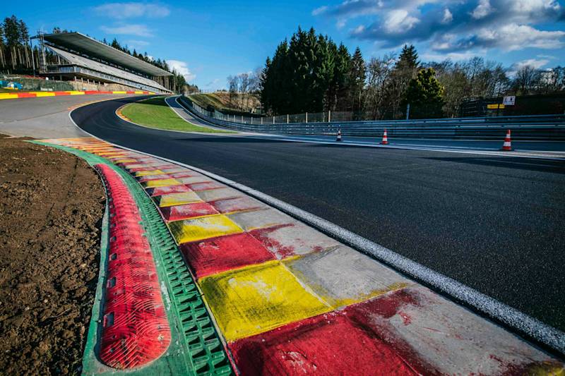 Porsche at racetrack corner with colorful curbs, trees, grandstand, and partly cloudy sky.