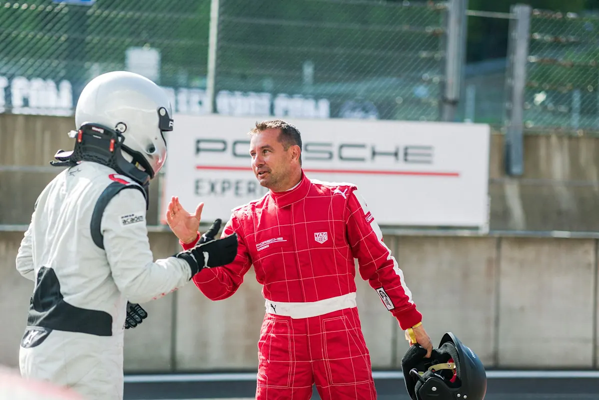 Rennfahrer in Anzügen und Helmen schütteln sich am Streckenrand die Hände, im Hintergrund ein Porsche-Schild.