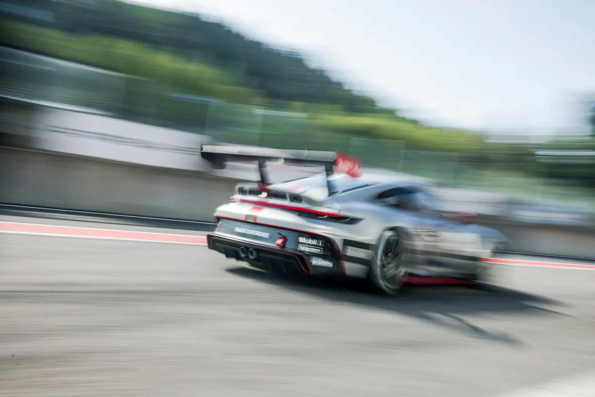 Verschwommener Porsche-Rennwagen rast auf der Strecke, dynamische Bewegung, Bäume im Hintergrund.