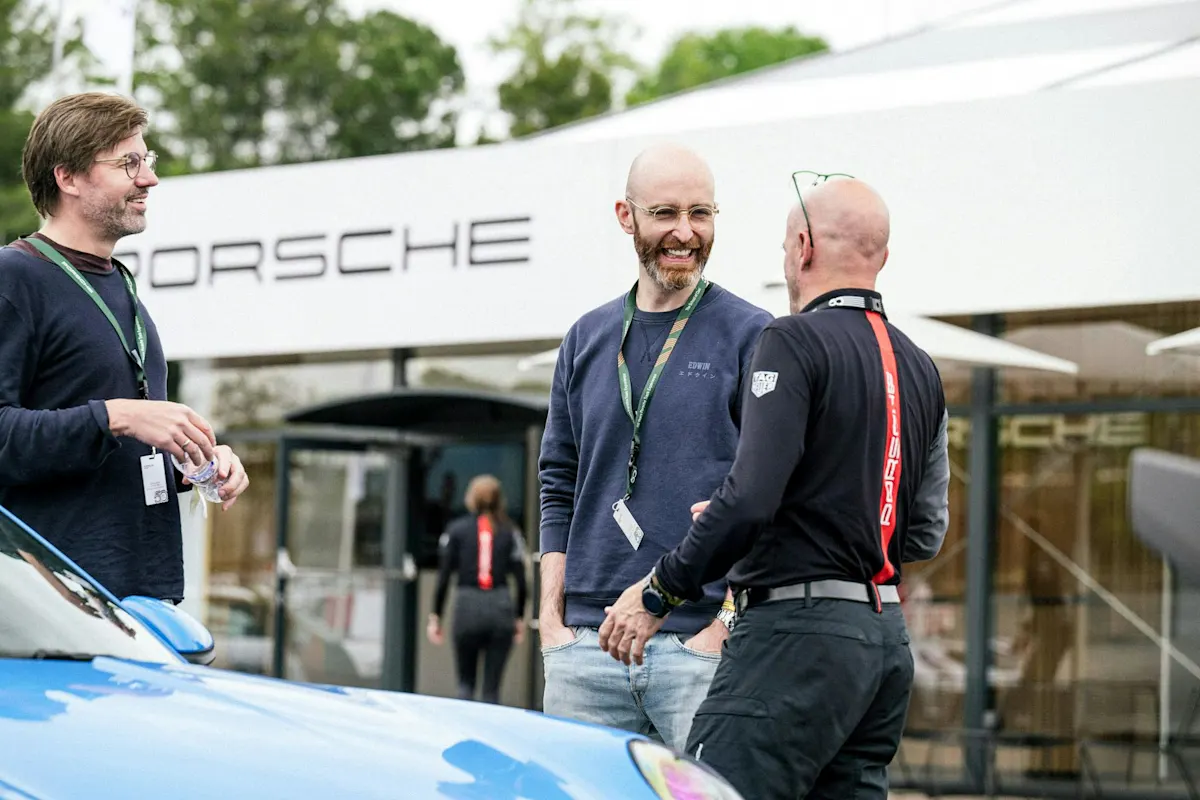 Drei Männer lächeln in der Nähe eines Porsches bei einer Outdoor-Veranstaltung, im Hintergrund Bäume und ein Porsche-Schild.