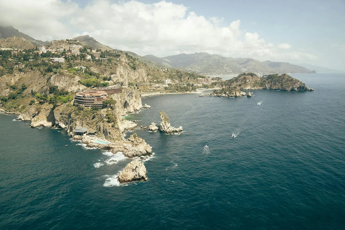 Aerial view of rocky coastline with buildings, green hills, boats at sea under cloudy sky.