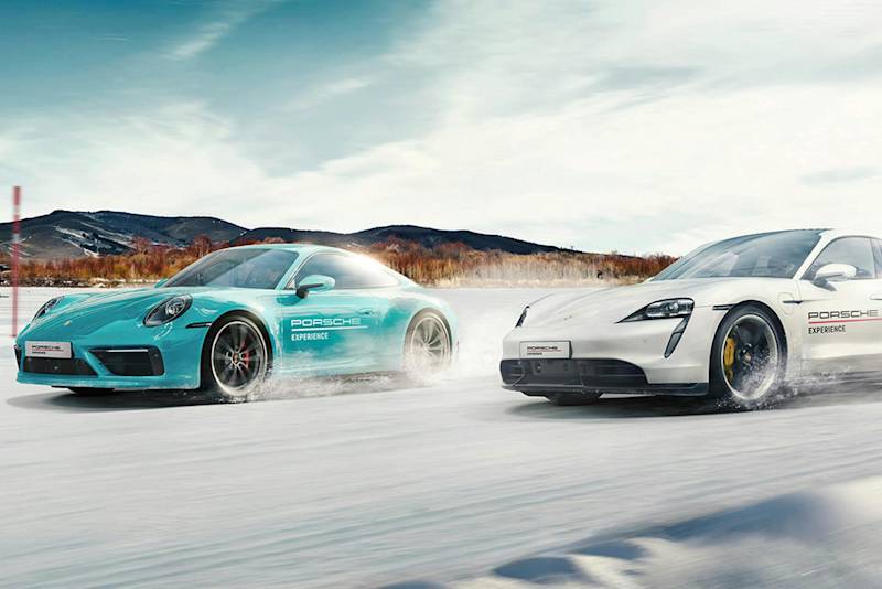 Porsche-Auto-Duo rast auf verschneiter Straße, blau links und weiß rechts, im Hintergrund schneebedeckte Berge und blauer Himmel.