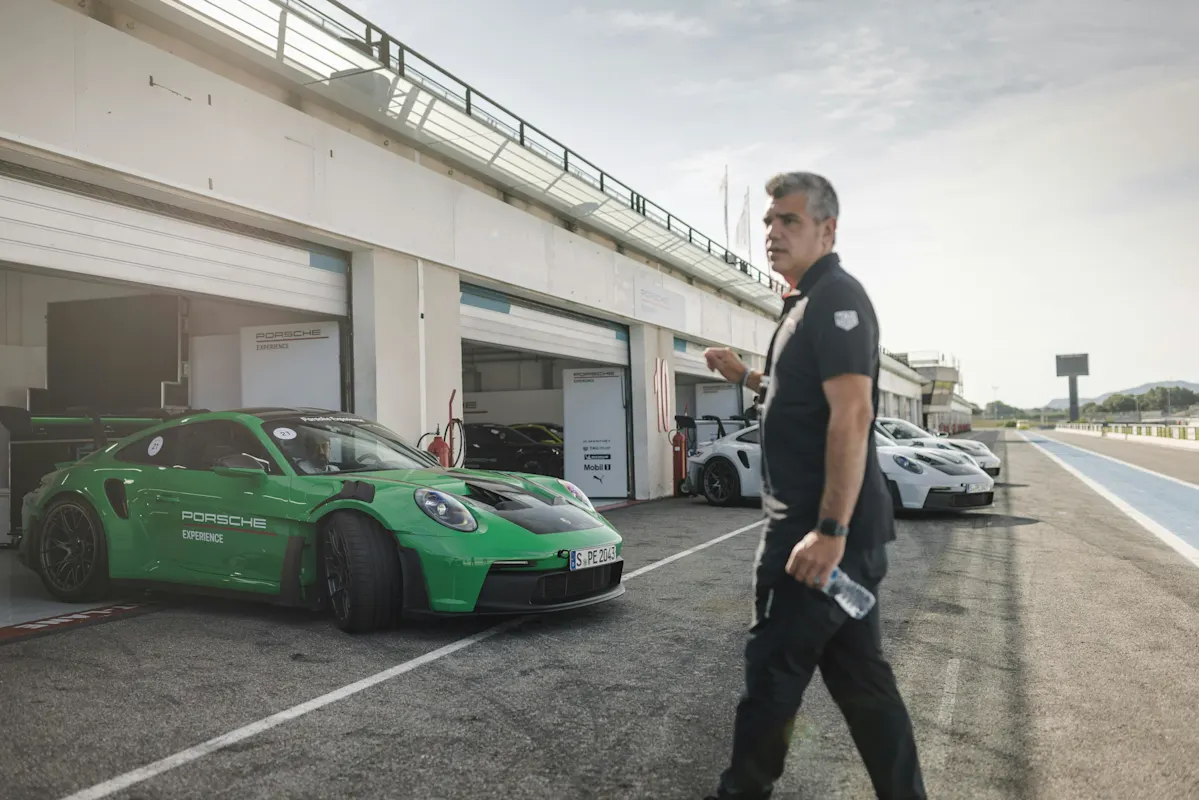 Eine Person geht an einem grünen Porsche in einer Rennstrecken-Garage vorbei, mit anderen Autos in der Nähe.