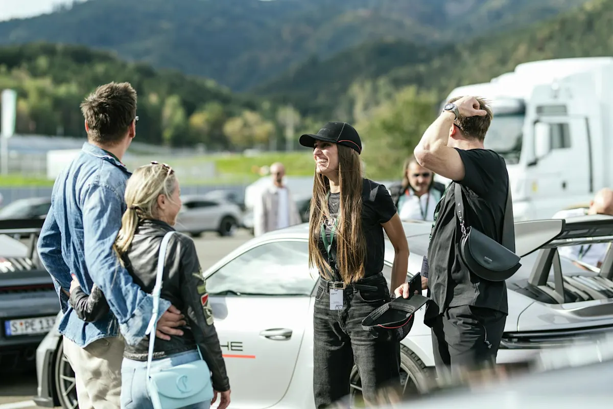 Menschen plaudern in sonnigem Außenbereich neben einem weißen Porsche mit Bergen im Hintergrund.