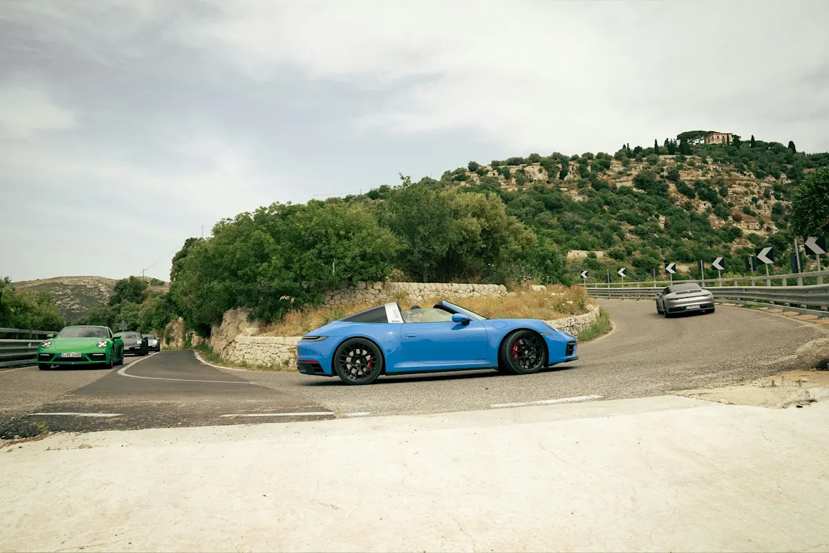 Blue Porsche convertible on winding road with green hills, followed by cars.