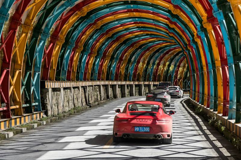 Ein roter Porsche Sportwagen führt eine Reihe durch einen farbenfrohen Metalltunnel, in dem Sonnenlicht gestreifte Schatten wirft.