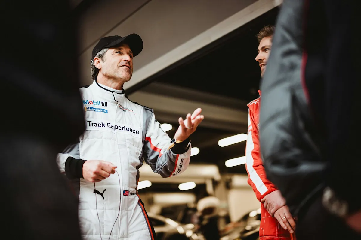 Man in a racing suit gestures and talks in a garage, people around him, including one in a red suit.
