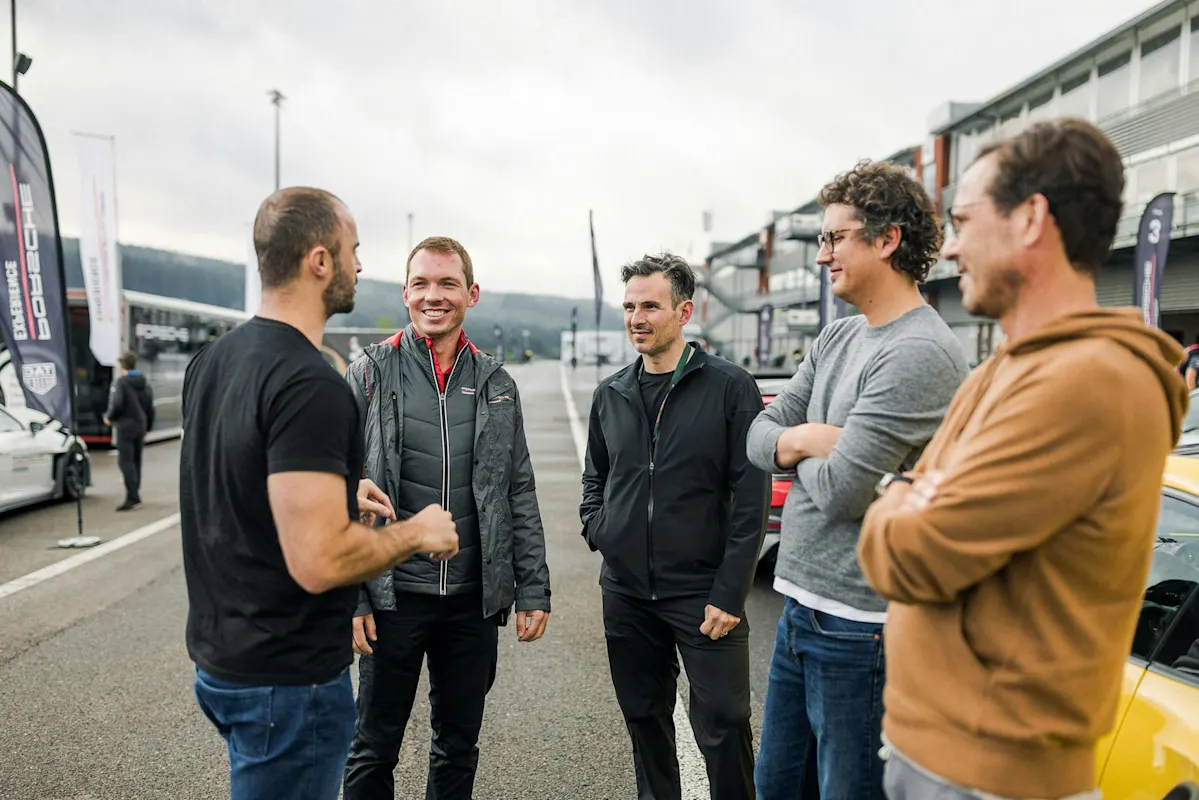 Fünf Männer unterhalten sich in einem Fahrerlager in der Nähe eines Porsche-Autos unter bewölktem Himmel.