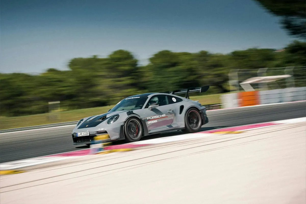 Silberner Porsche Sportwagen rast auf der Rennstrecke, unscharfer Hintergrund, entfernte Bäume und Barrieren.