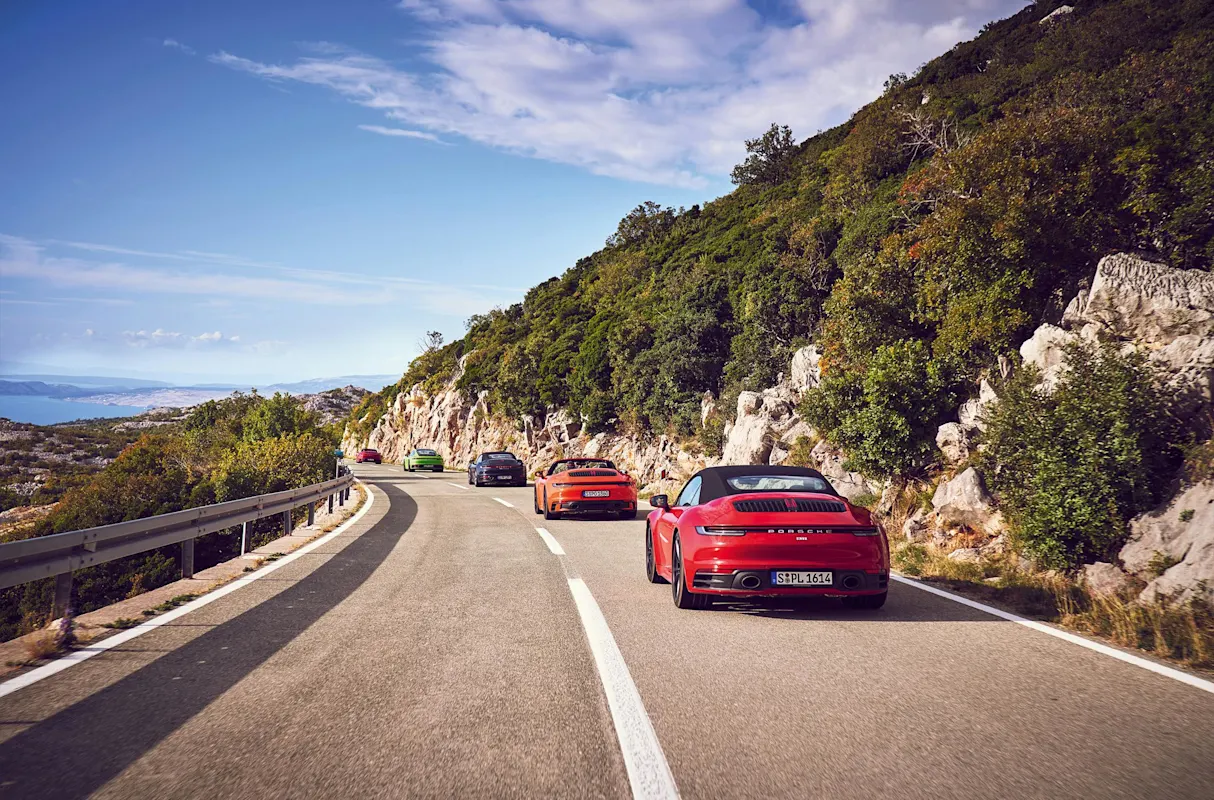 Porsche Sportwagen fährt auf malerischer Küstenstraße mit felsigen Klippen und üppigem Grün unter blauem Himmel.