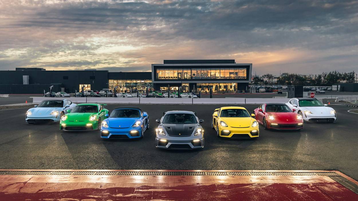 Eine Reihe von sieben Porsche-Sportwagen in verschiedenen Farben auf einer Rennstrecke, mit einem modernen Gebäude gegen einen Abendhimmel.