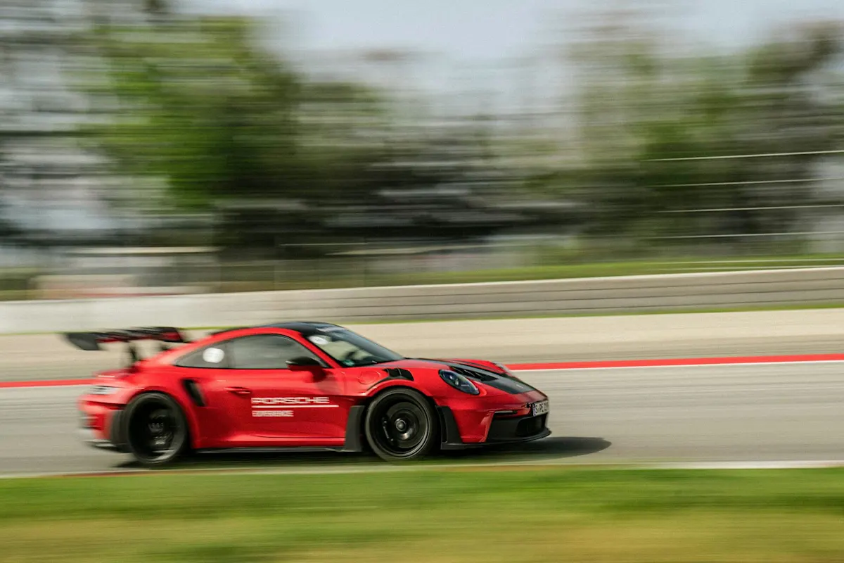 Ein roter Porsche rast auf der Rennstrecke mit verschwommenem Effekt, Bäumen und Barrikaden im Hintergrund.