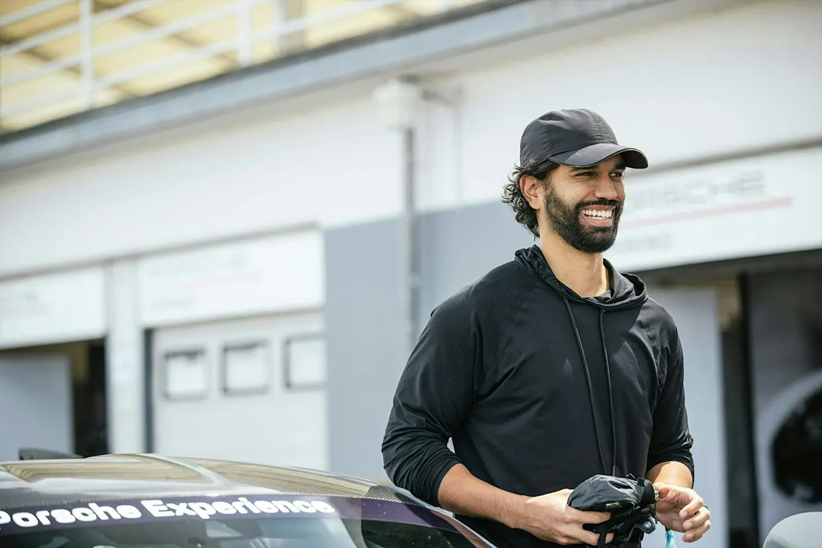 Mann in schwarzer Kapuzenjacke und Kappe lächelt neben einem Porsche Experience Auto.