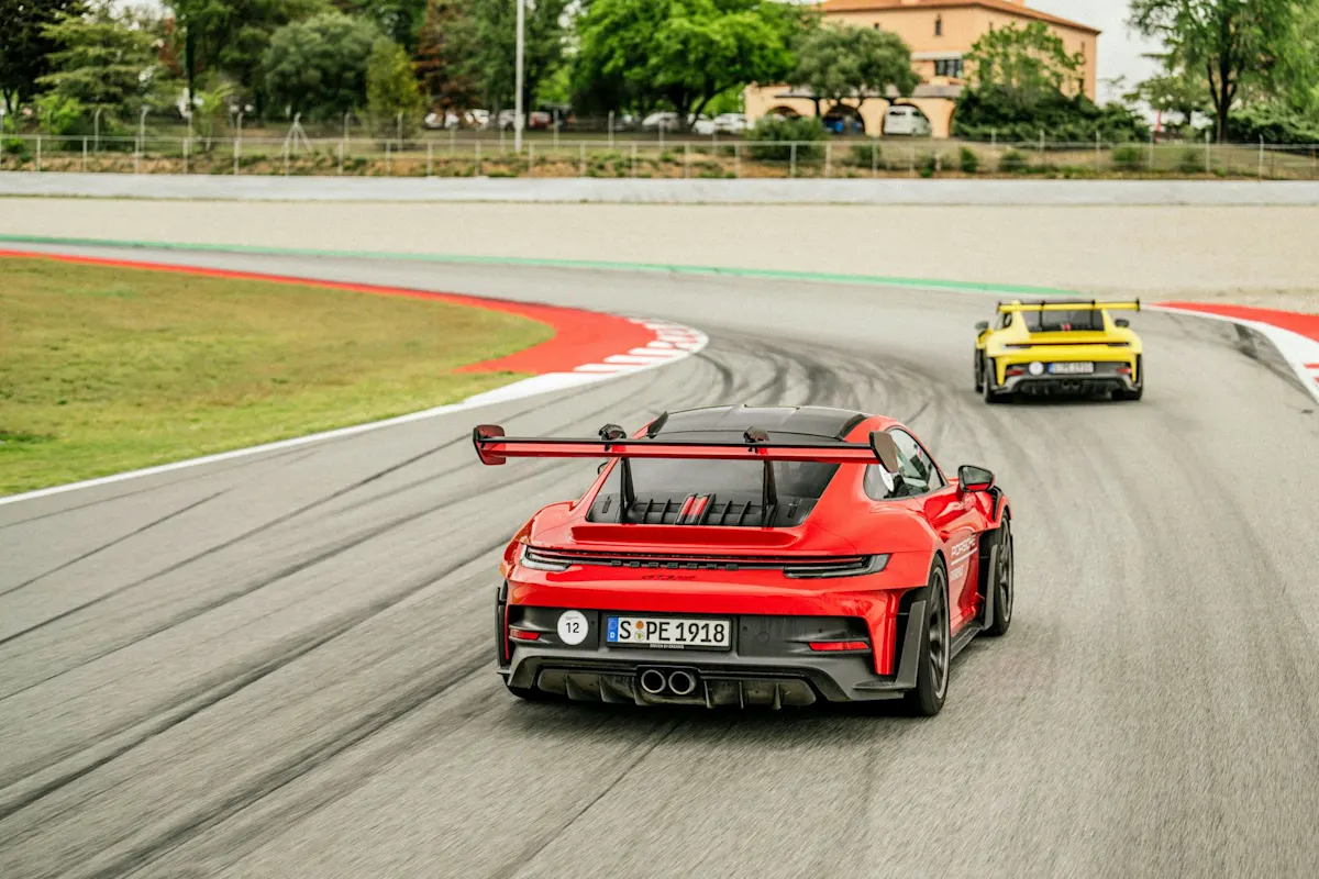 Rote und gelbe Porsche Sportwagen rasen auf einer Rennstrecke mit bunten Randsteinen, Grünflächen und einem entfernten Gebäude.