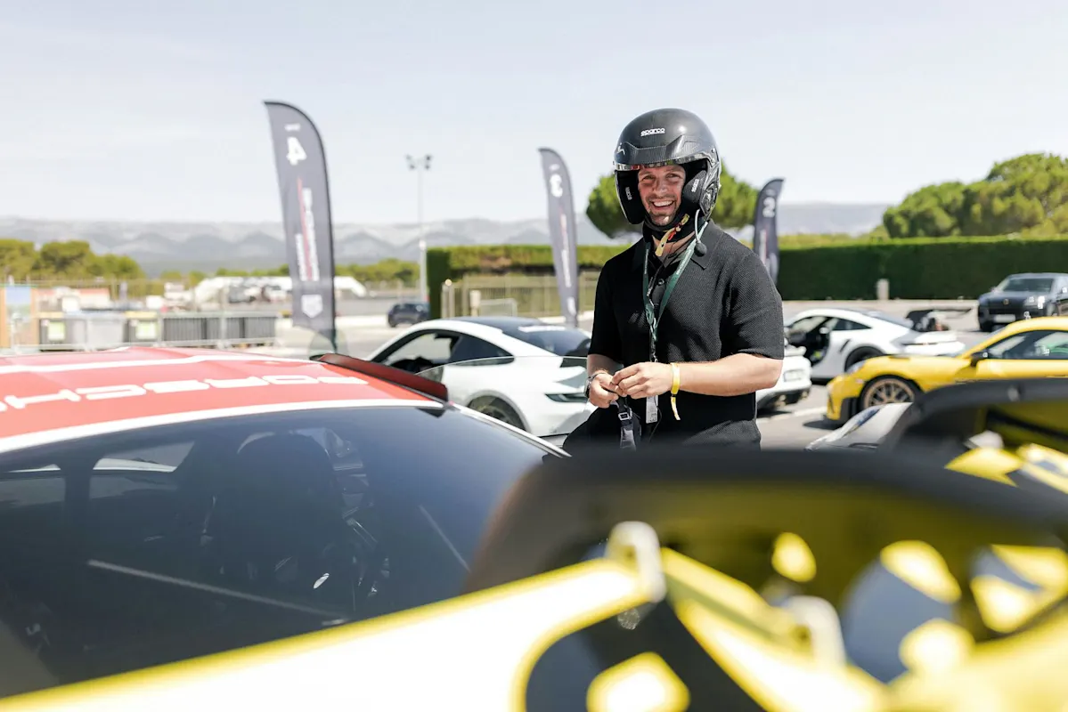 Person in Helm neben einem Porsche Sportwagen an sonnigem Tag, mit anderen Autos und Bergen.