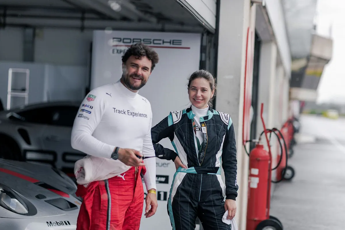 Two individuals in racing outfits smile near a Porsche, next to a garage and a fire extinguisher.