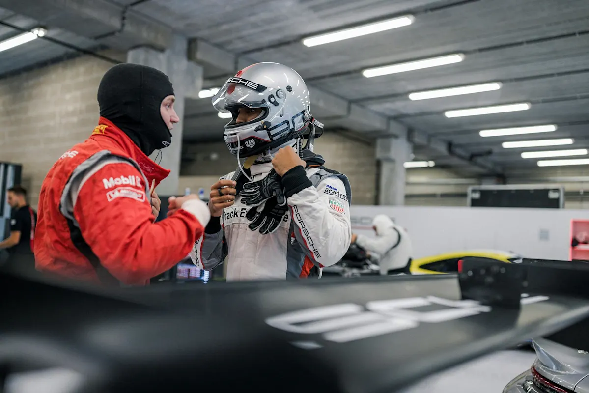 Race car driver in red suit with balaclava, adjusting helmet, next to car wing.