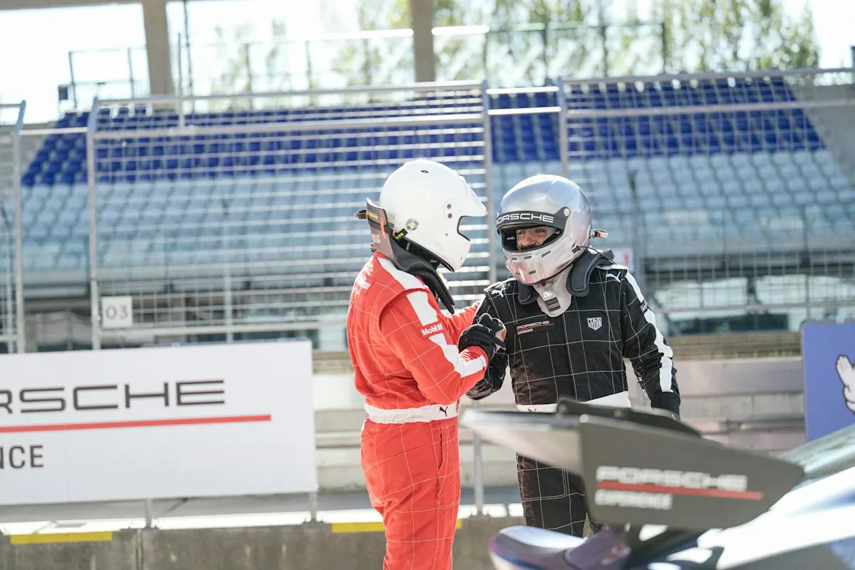 Race car drivers in helmets near a Porsche race car at a track, with seating in the background.