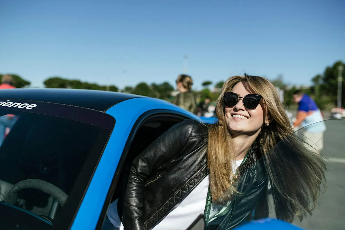 A woman with long hair and sunglasses smiles out of blue Porsche car window under clear sky.