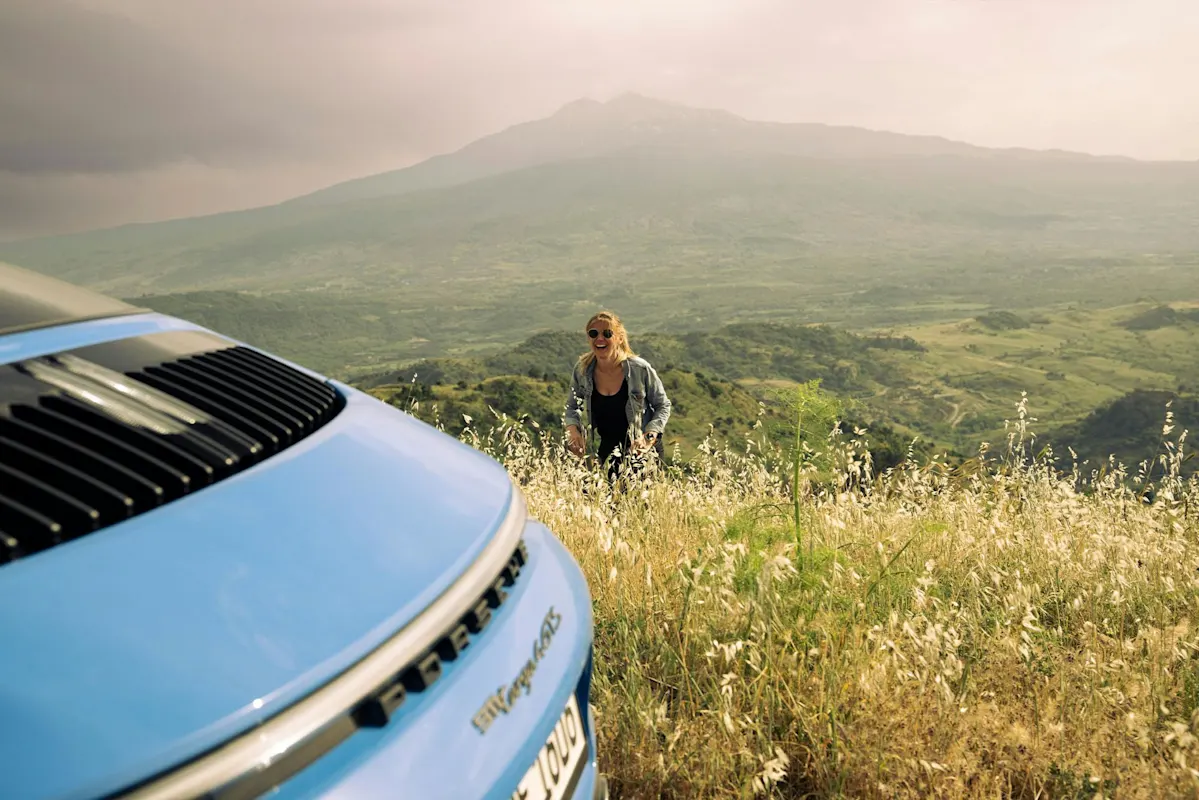 Person in field with mountain backdrop, blue Porsche parked in front.