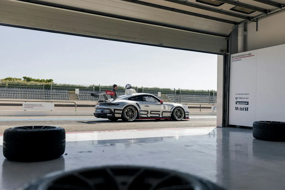 Silver Porsche race car with number 5 in garage on racetrack, person nearby.