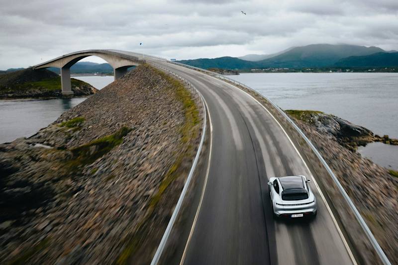 A Porsche sports car drives on a coastal highway with an arch bridge, rocky terrain, water, and mountains in the background.