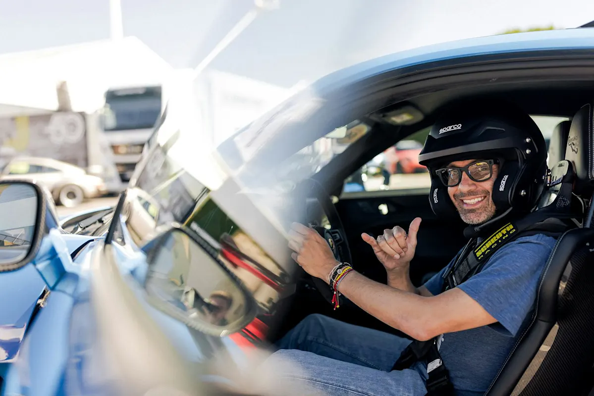 Person in einem blauen Porsche mit Helm und Sonnenbrille gibt Daumen hoch. Hintergrund mit Fahrzeugen und Gebäuden.