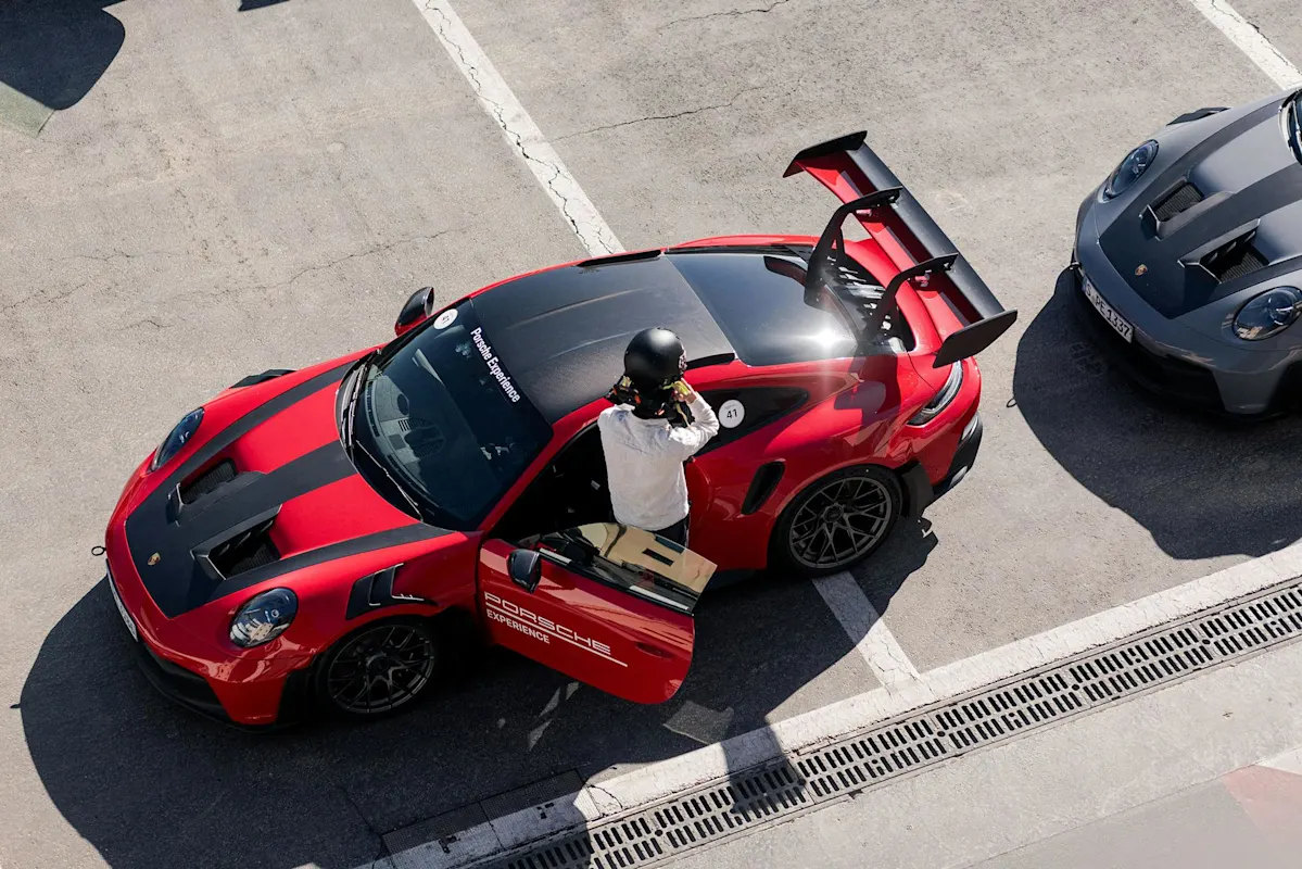Luftaufnahme eines roten Porsche-Sportwagens mit schwarzer Motorhaube und Spoiler neben grauem Auto geparkt. Person mit Helm steht dabei.