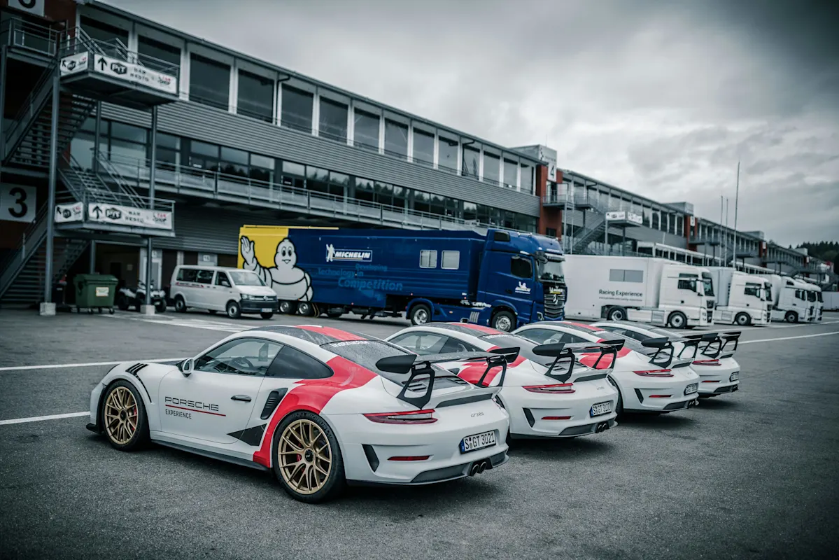 Three white Porsche race cars with red accents parked in a row at a racetrack, trailers and trucks nearby under a cloudy sky.