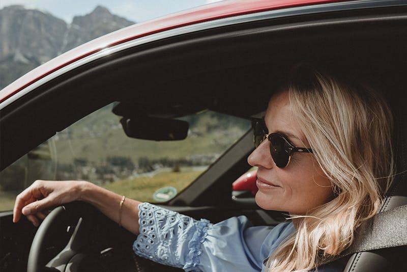 Frau in Sonnenbrille fährt roten Porsche durch malerische Berglandschaft.