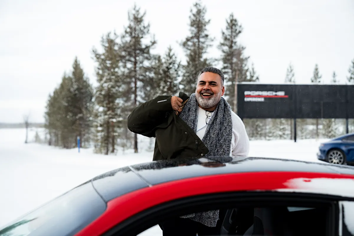 A man stands by a car in snowy area with trees and Porsche Experience sign in background.