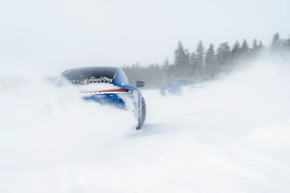 Blue Porsche 911 drifts on snowy track, flying snow partially obscures the car. Pine trees in the background.