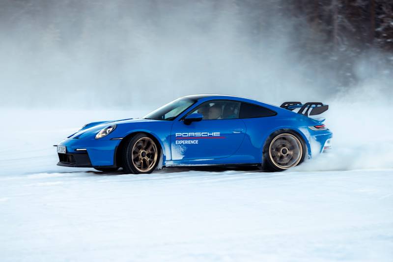 A blue Porsche speeds on snowy surface, kicking up ice and snow.