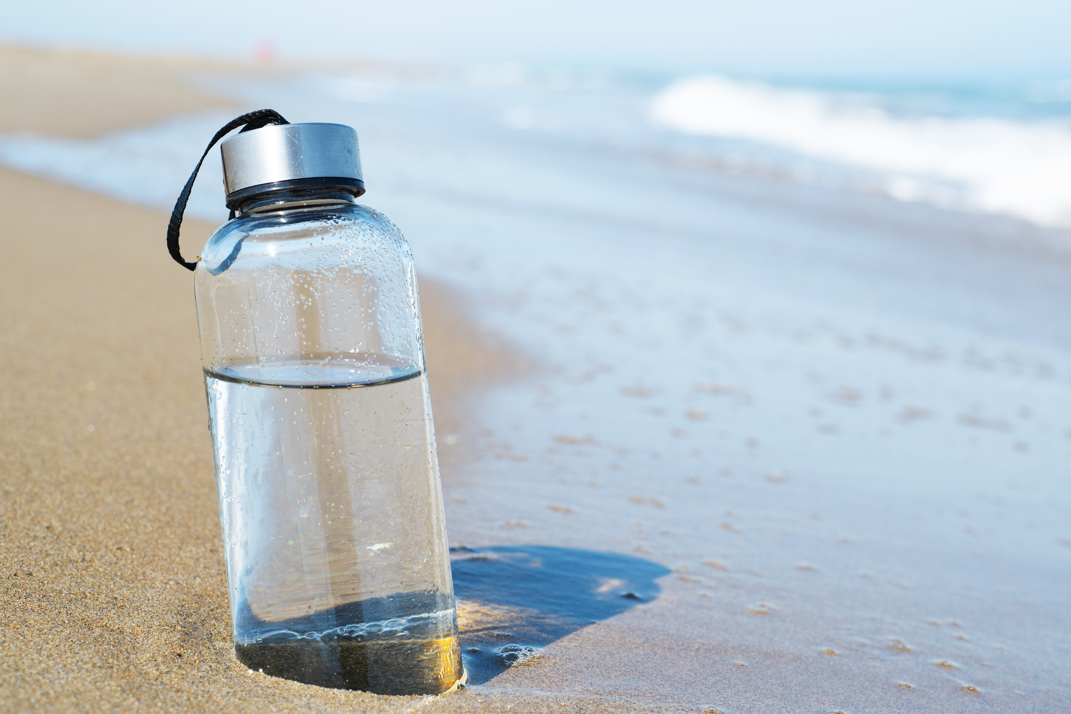 A glass reusable water bottle on a beach for Plastic Free July.