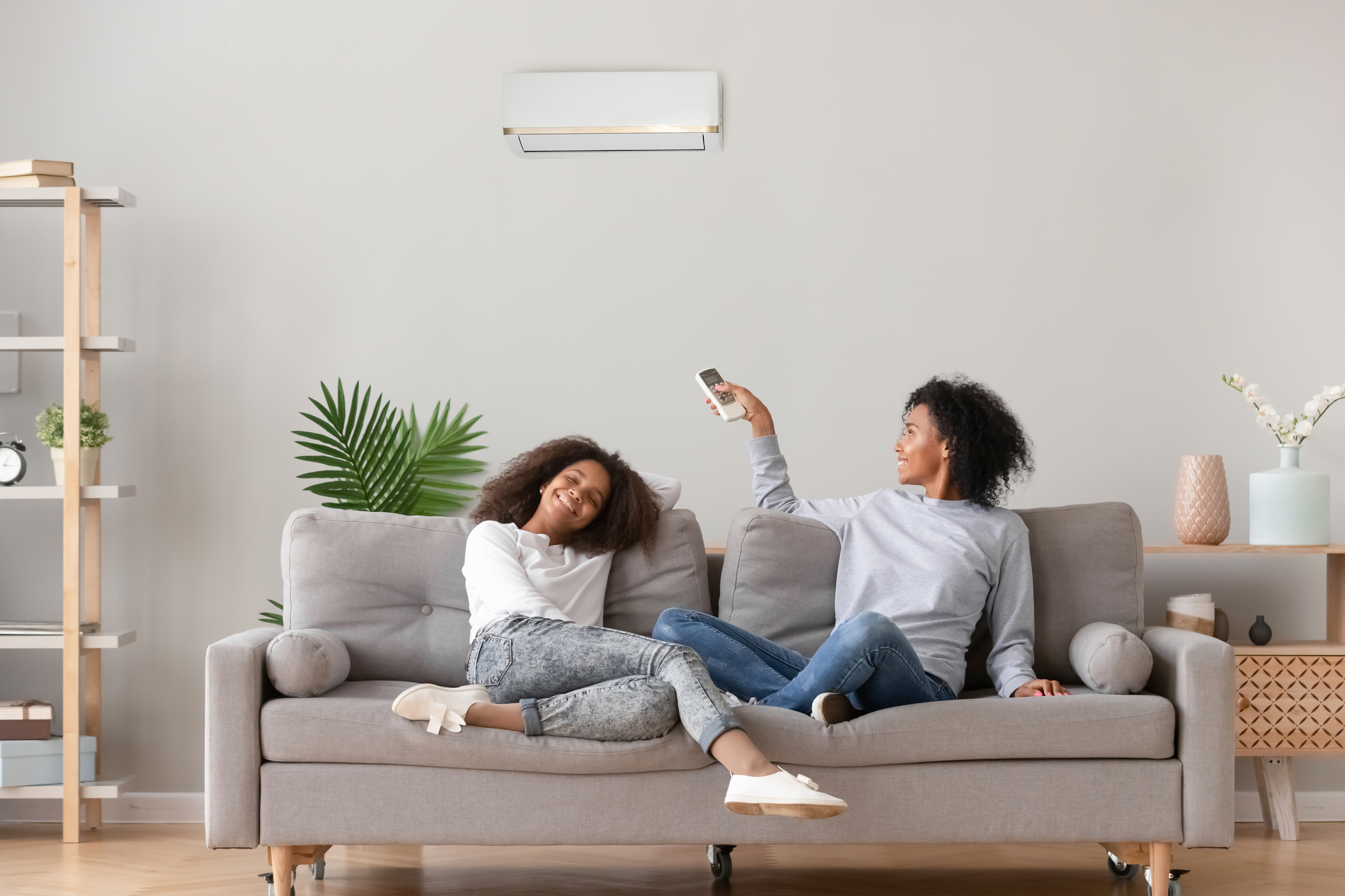 Two women smiling sitting on a couch in their home, adjusting their air conditioning unit with a remote.