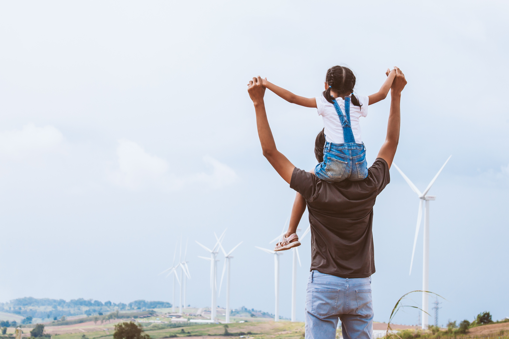 A daughter sits on her father's shoulders looking out over a wind farm
