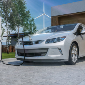 An electric car charging in a driveway with a wind turbine in the background.