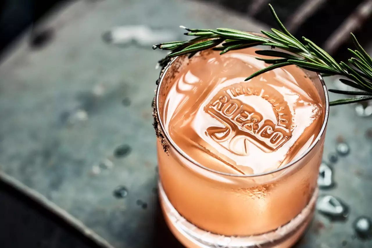 A Roe & Co cocktail with a branded ice cube and rosemary garnish on a spiced rim glass, served on a metal surface.