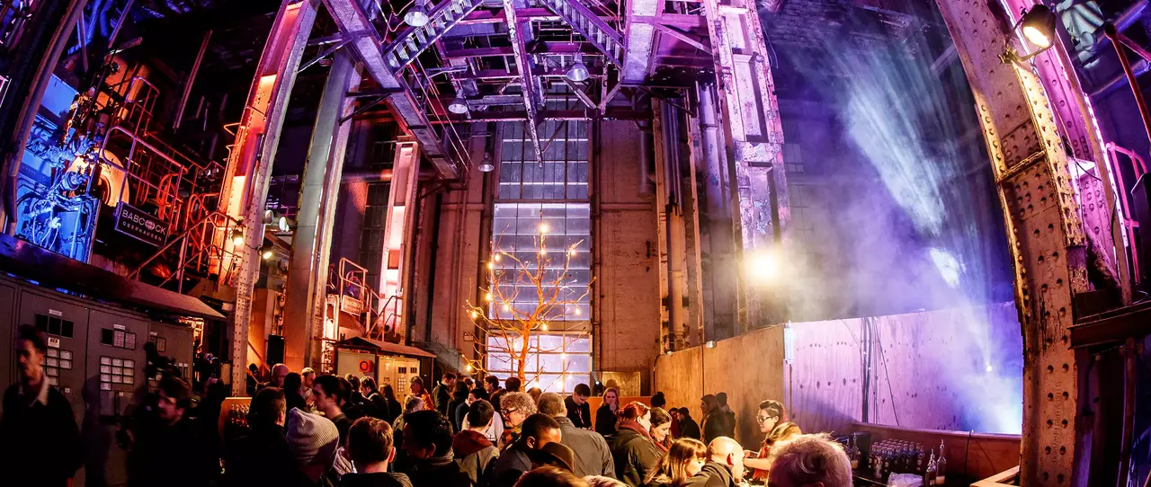 Crowd gathers inside an industrial venue with exposed steel beams, colourful stage lighting, and an illuminated tree, during a lively event.