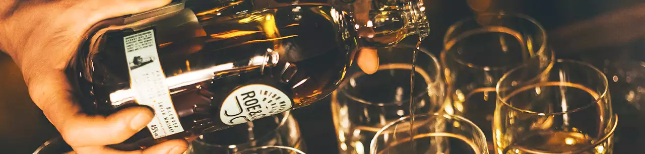 Bartender pours Roe & Co Blended Irish whiskey into multiple clear glasses at a bar, close-up of hands and bottle during drink preparation.