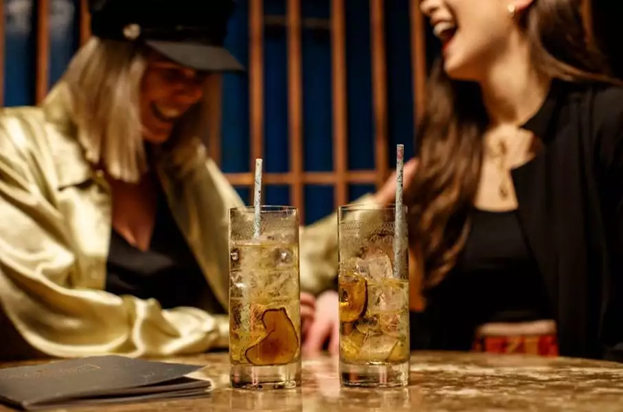 Two women laugh over cocktails garnished with dried citrus slices and paper straws at a warmly lit bar.