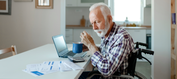 An elderly man in a wheelchair drinking coffee while working on a laptop at home.